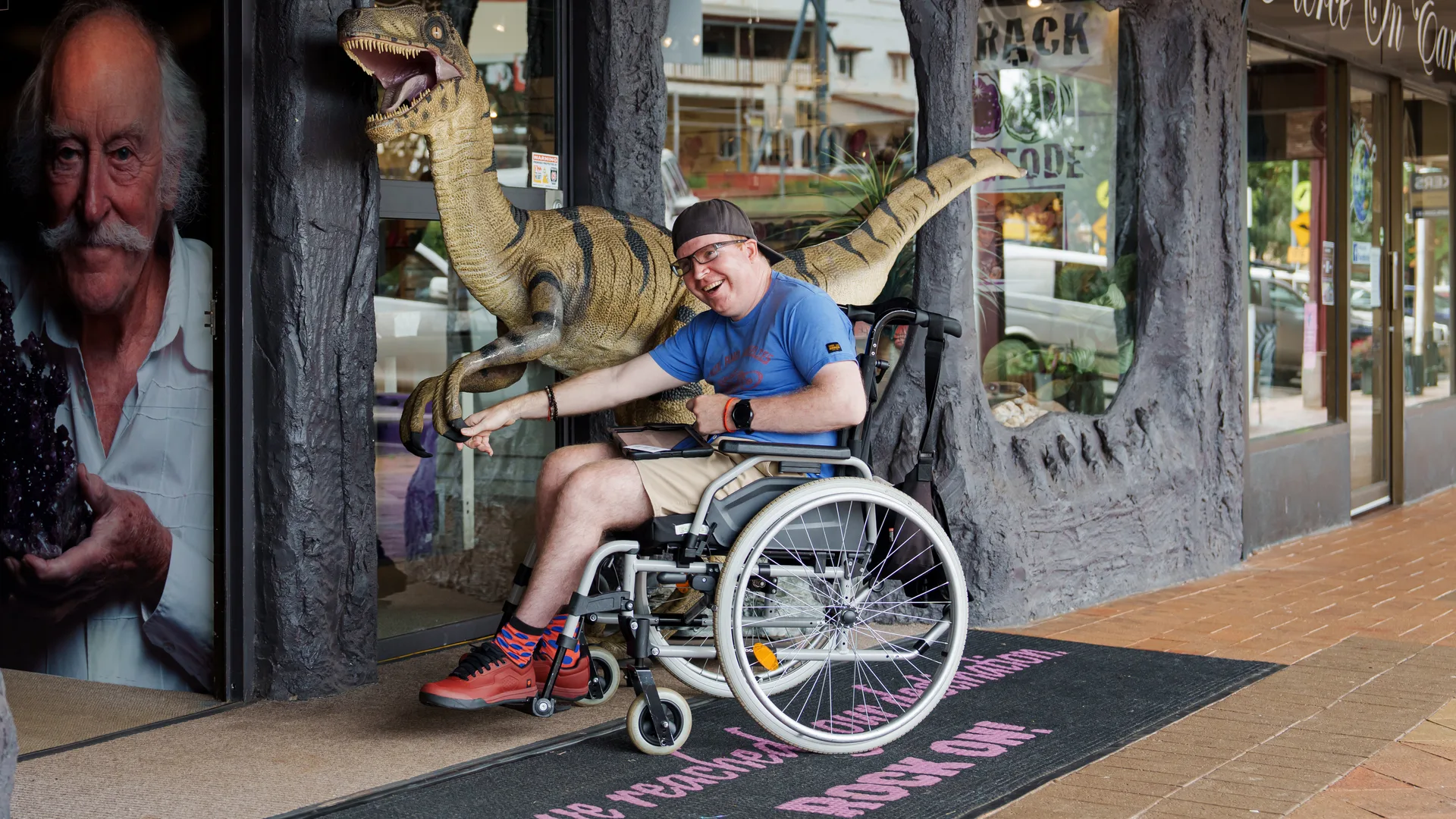 A visitor in a wheelchair at the Crystal Caves entrance, with a velociraptor model overhead